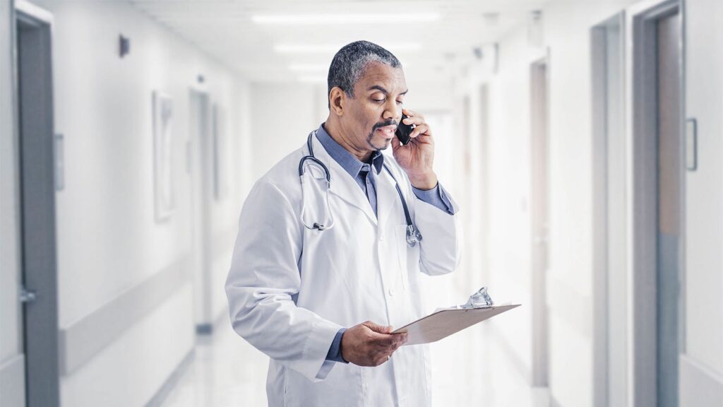 A photo of a mature male physician standing in a hospital hallway holding a clipboard and talking on the phone